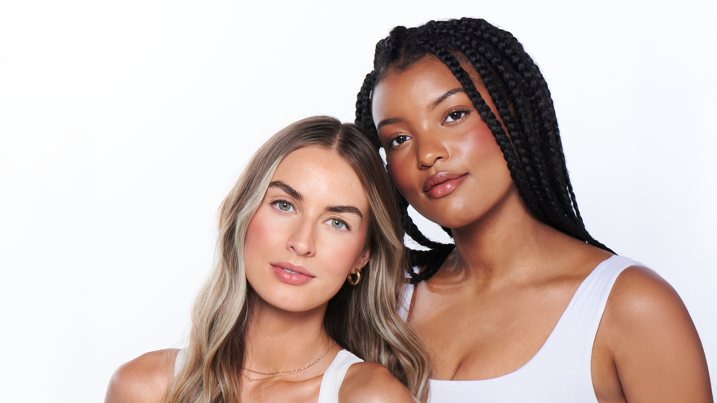 Two models side by side with natural casual makeup, one with blonde wavy hair, one with long box braids, both wearing white tops, soft natural light, clean white background