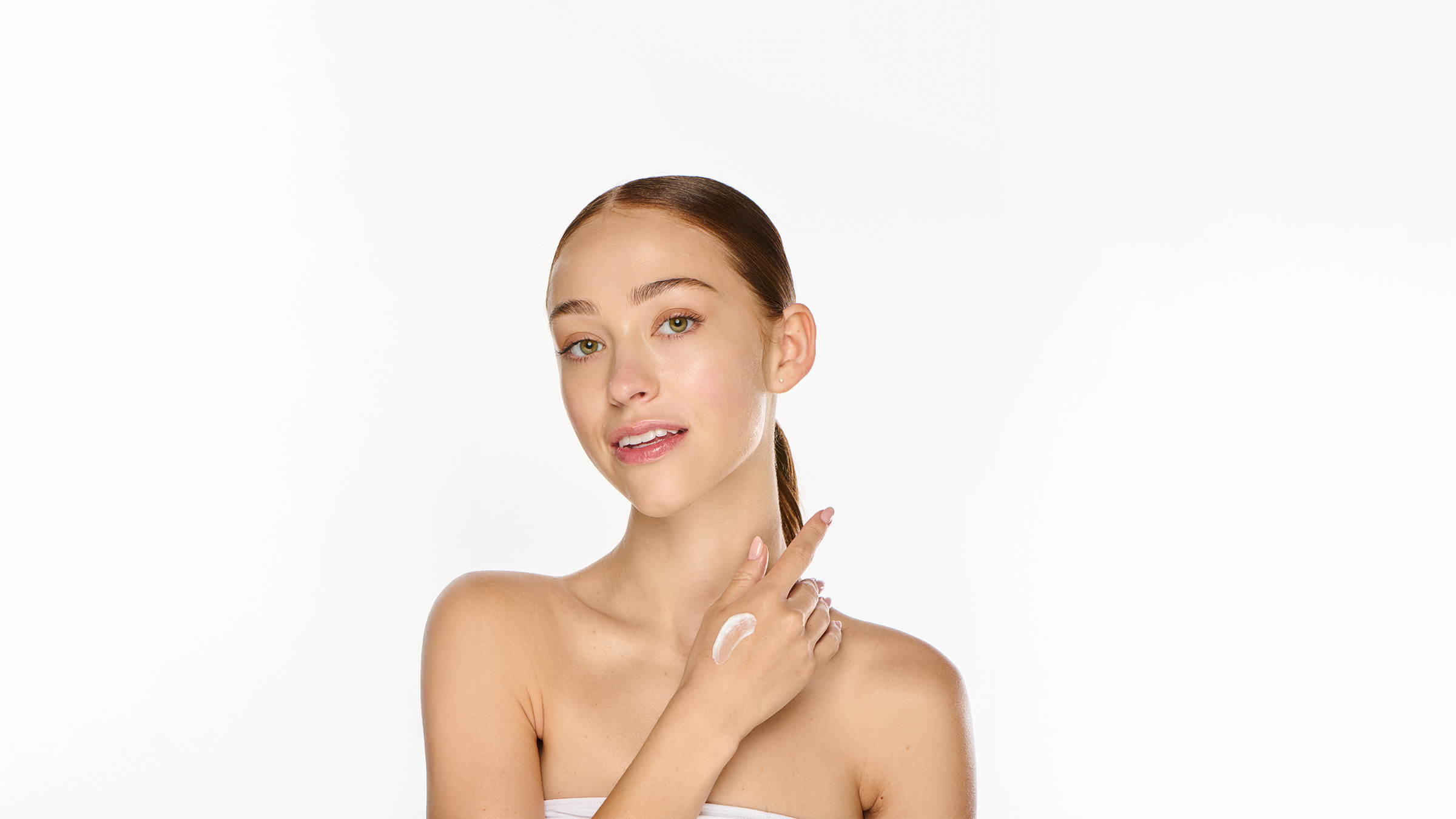 Young model with smooth glowing skin, brown hair pulled back, wearing a white strapless top, hand resting at her shoulder, soft natural light, clean white background