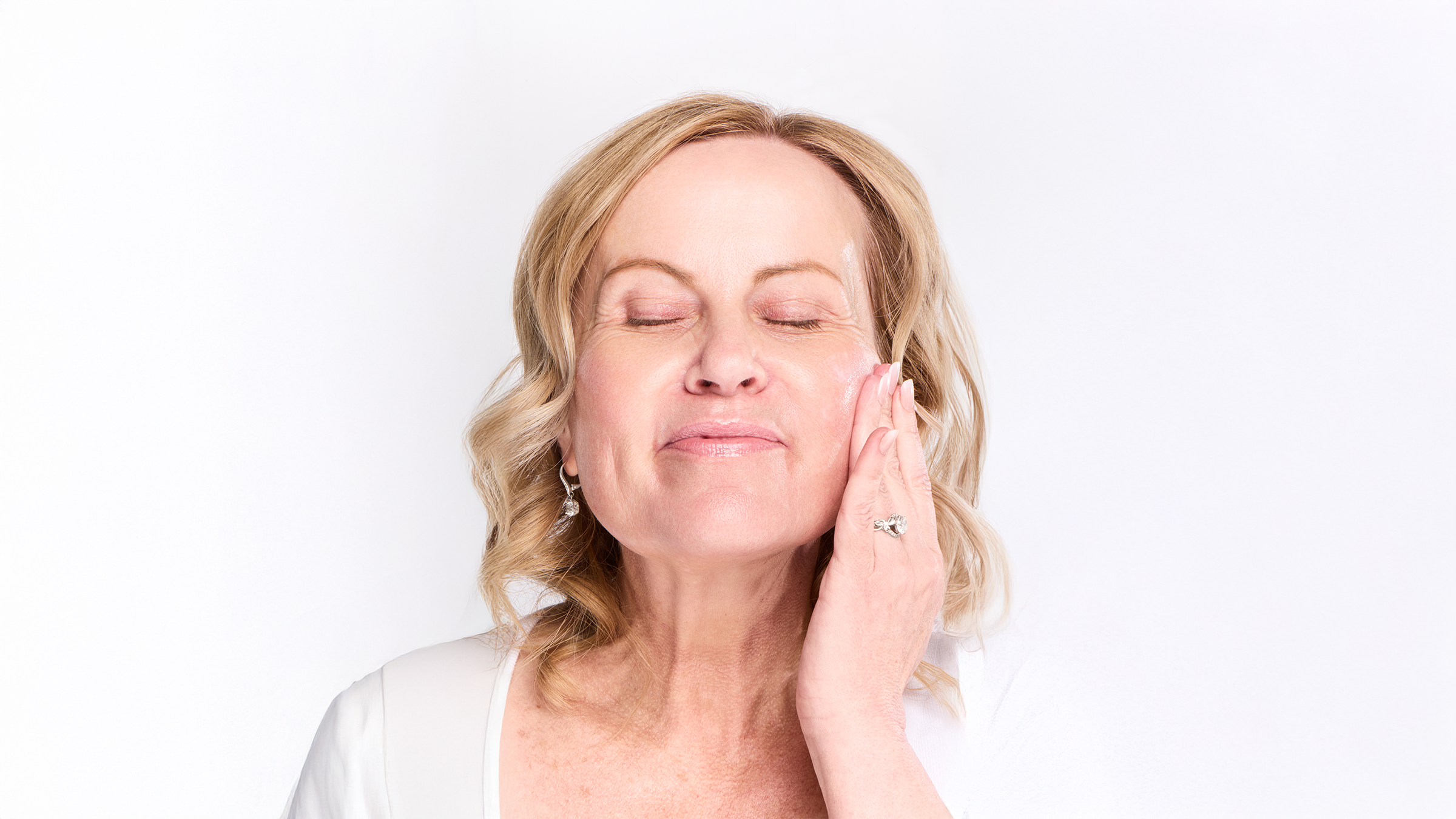 Older blonde woman with closed eyes and a soft smile, applying moisturizer to her cheek with her fingertips, wearing a white top, soft natural light, clean white background
