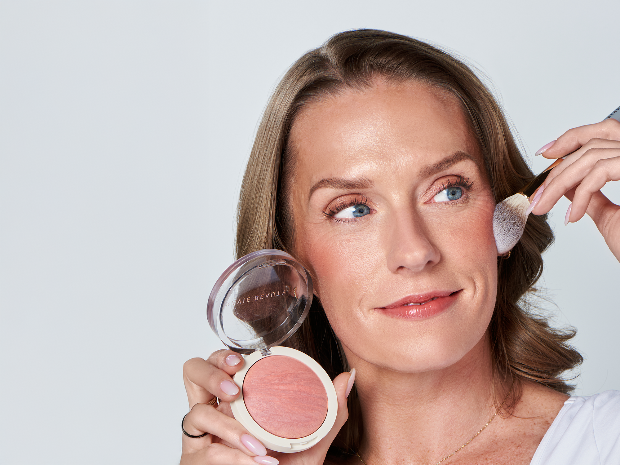Mature blonde model holding an open Ravie Beauty Endless Diffusion Baked Blush compact while a fluffy brush sweeps peachy pink blush across her cheekbone, soft natural light, clean white background