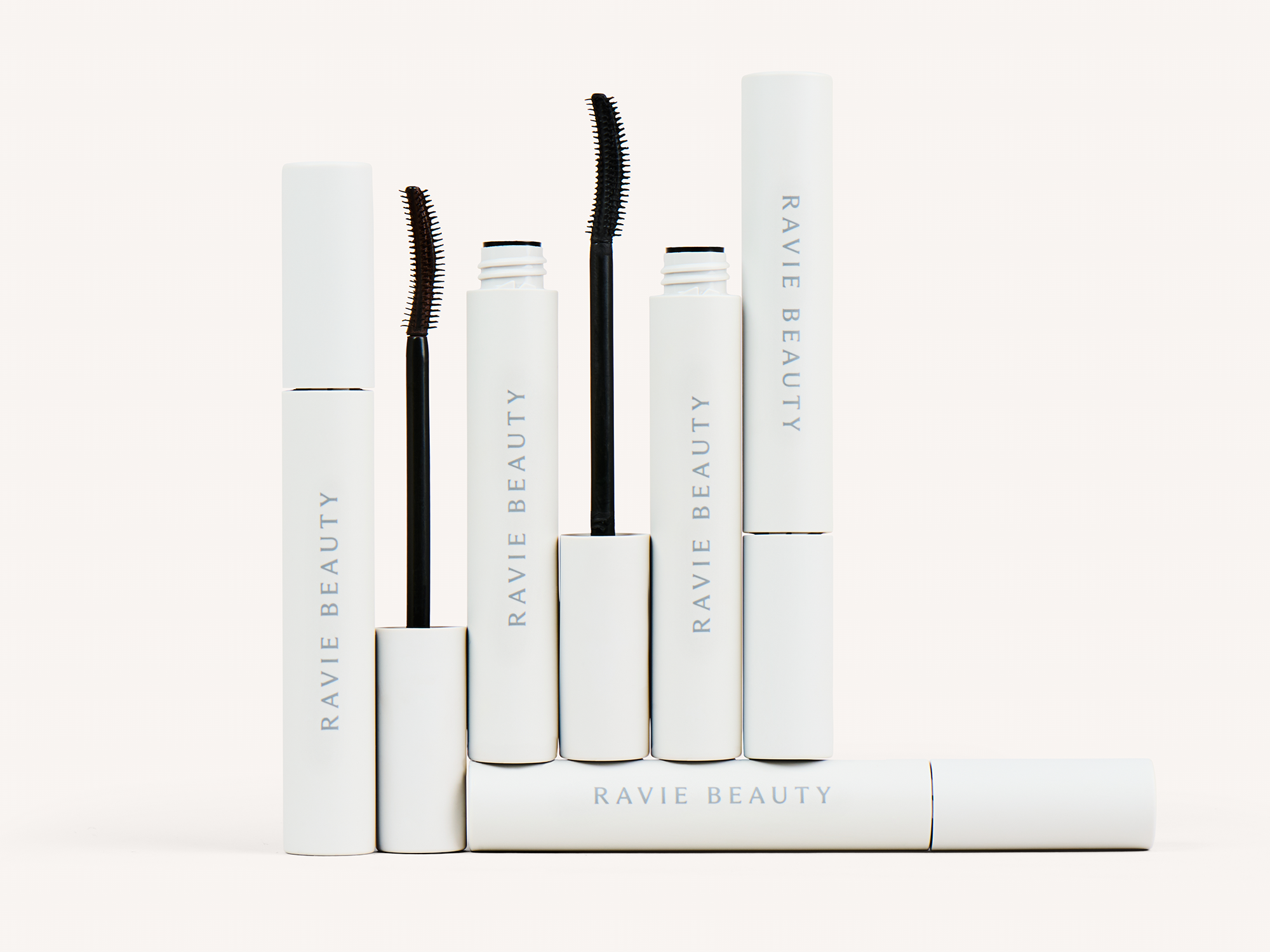 Five Ravie Beauty Effortless Lash mascara tubes arranged in a cluster on a soft cream background, two with wands extended to show the curved brush heads, soft natural light, minimal product styling