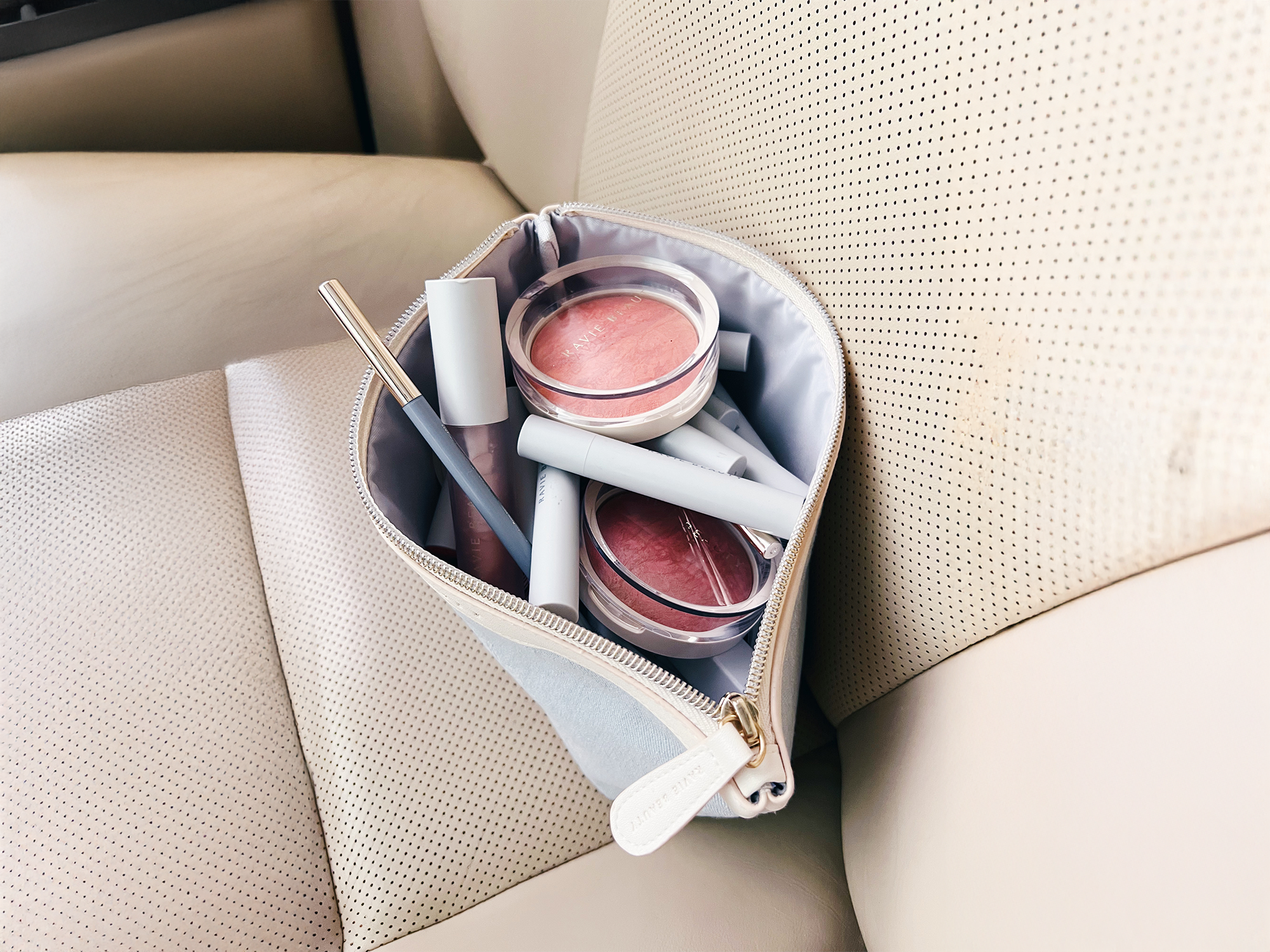 Open beige zip pouch on a cream perforated leather car seat containing Ravie Beauty Effortless Lips bullets, Endless Diffusion Baked Blush compacts, and a lip product, soft natural daylight, lifestyle still life