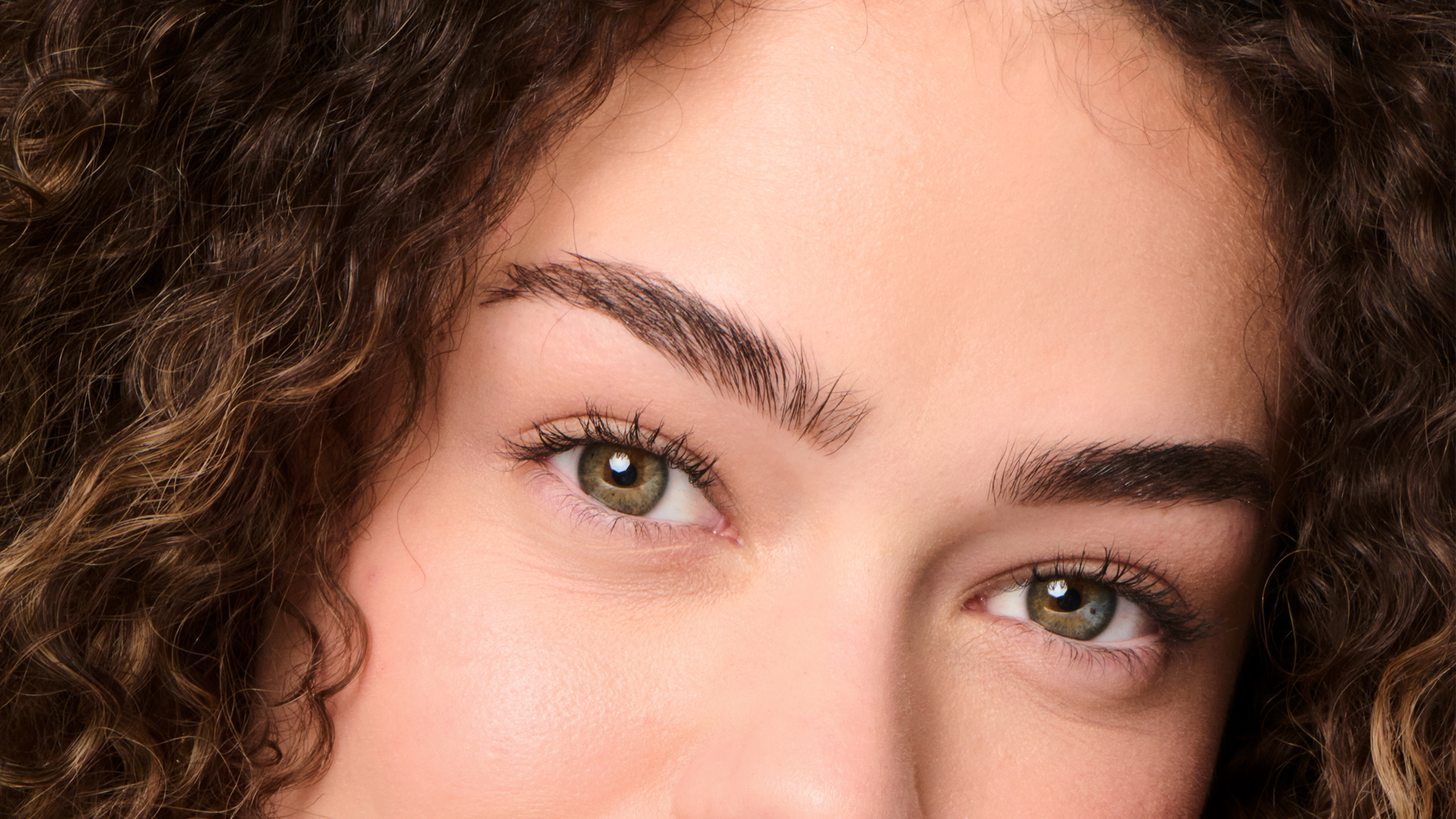 Close-up of green eyes framed by full brown brows and curly auburn hair, editorial detail shot in soft natural light