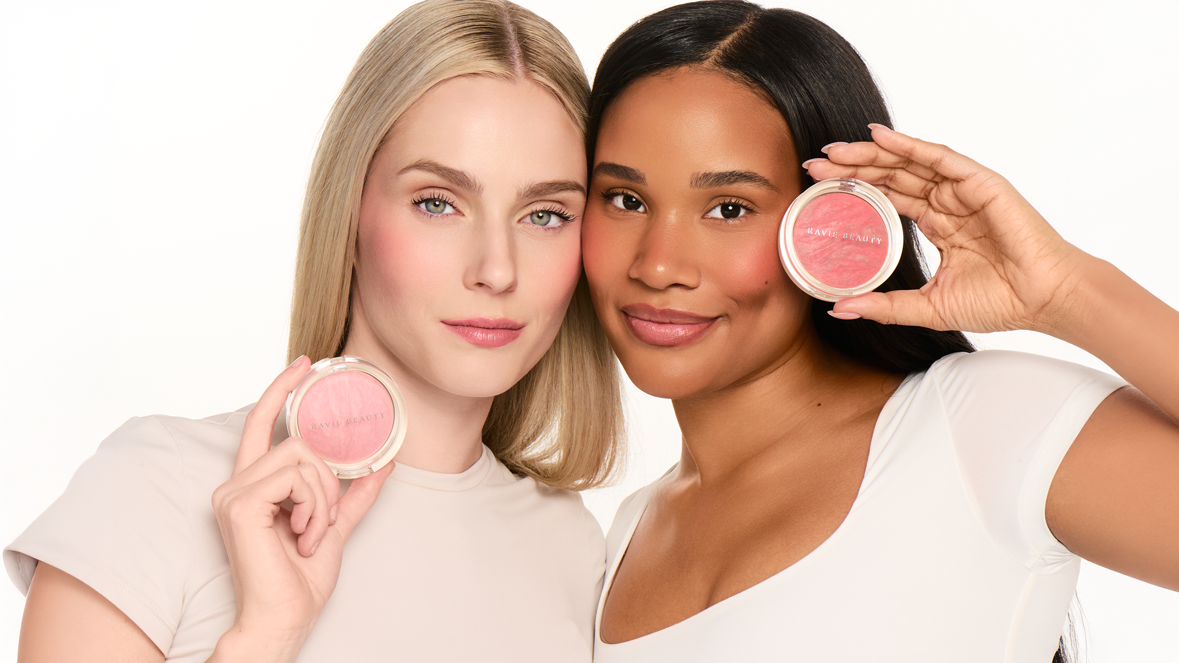 Two smiling models holding open Ravie Beauty Endless Diffusion Baked Blush compacts showing peachy pink shades, one with blonde hair, one with dark hair, soft natural light, clean white background