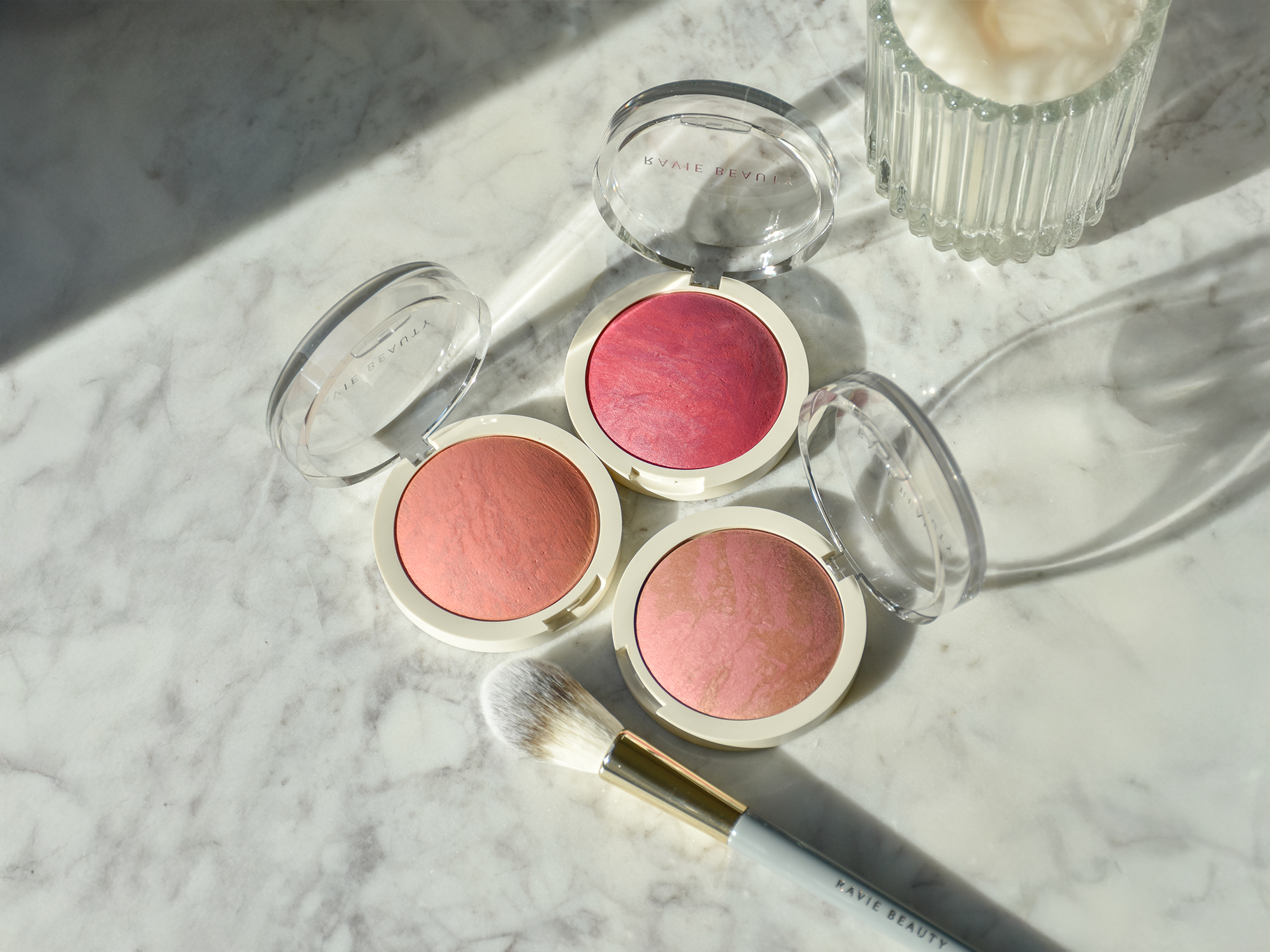 Three open Ravie Beauty Endless Diffusion Baked Blush compacts in peachy pink and rose shades arranged on a marble surface with a fluffy blush brush, soft natural light, editorial flatlay styling
