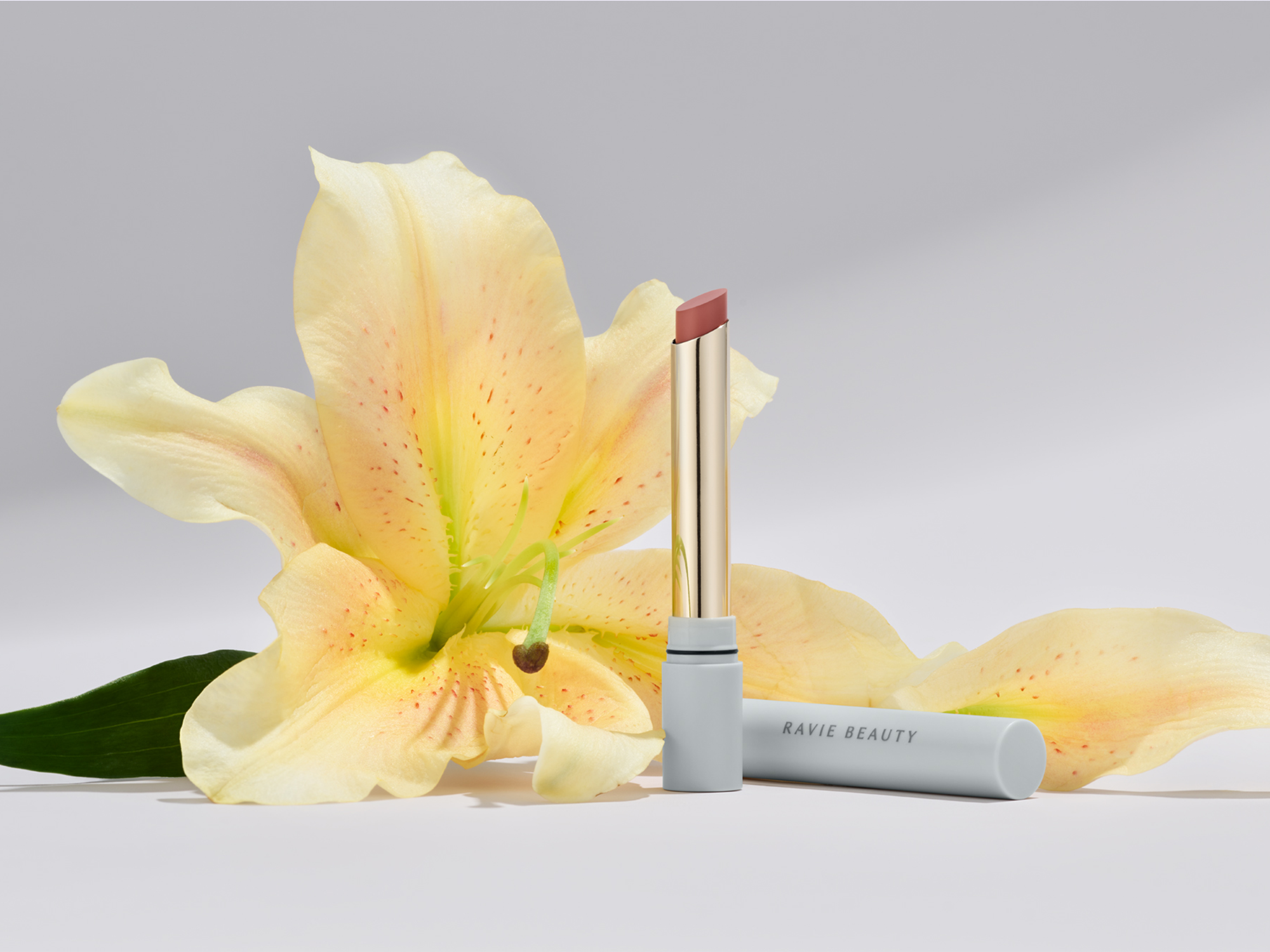 Ravie Beauty Effortless Lips bullet open in a warm peachy nude shade propped against a soft yellow lily blossom with fallen petals on a pale grey surface, soft natural light, editorial styling