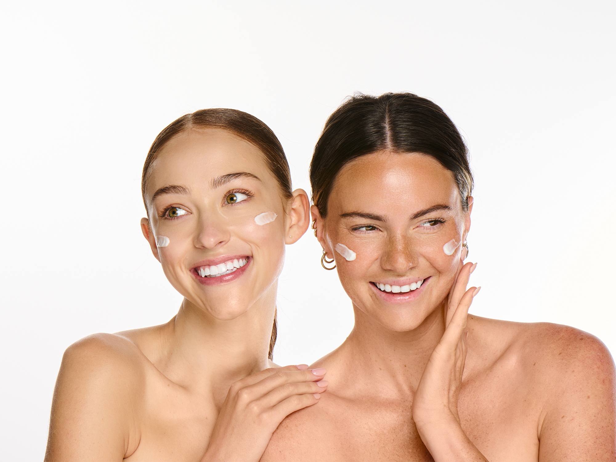 Two smiling models with bare glowing skin and dabs of Ravie Beauty moisturizer on their cheeks and foreheads, soft natural light, clean white background