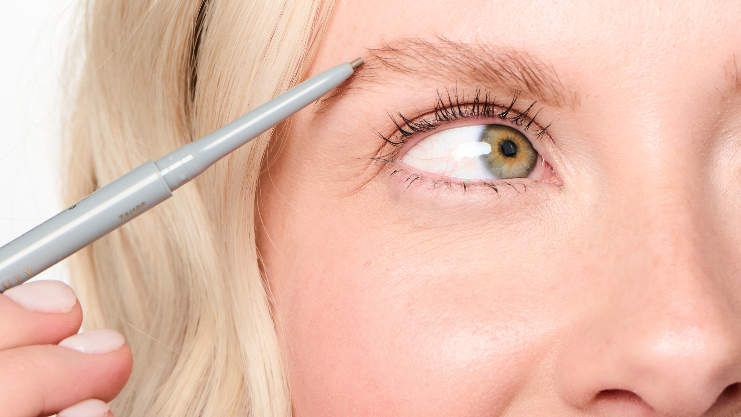 Close-up of a blonde model's brow being filled with a Ravie Beauty Easy Everyday Brow Pencil, hand visible holding the pencil, soft natural light, clean background