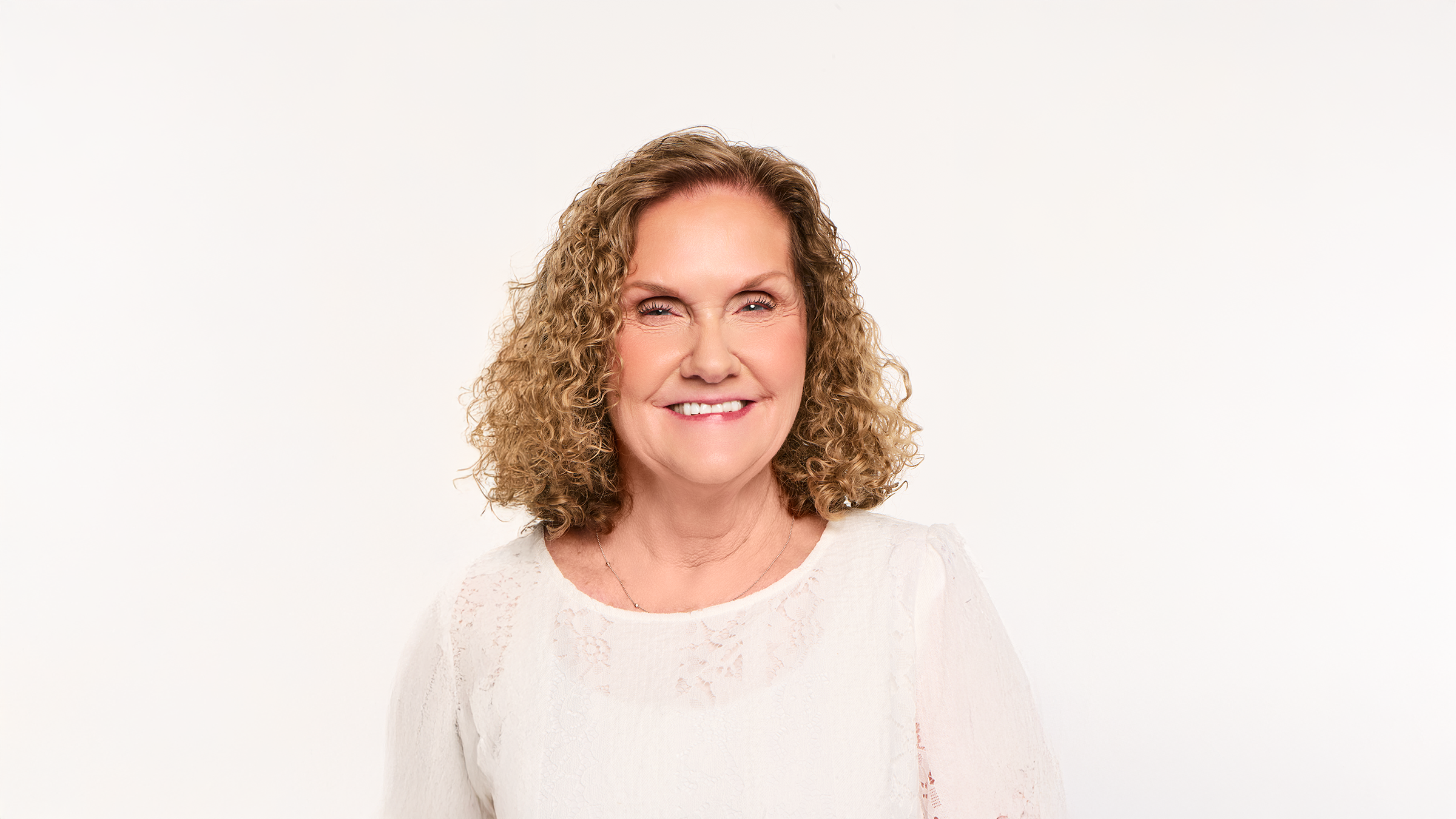 Smiling woman with curly blonde hair and natural makeup wearing a white embroidered top, soft studio light, clean white background