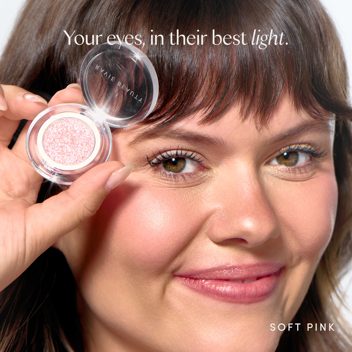 Your eyes, in their best light. A model wearing it, holding the compact. Ravie Beauty Softlight™ Glistening Eye Topper in Soft Pink 
