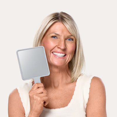 A smiling woman holding The Hand Mirror, mirror side out, beside her face. Ravie Beauty The Hand Mirror